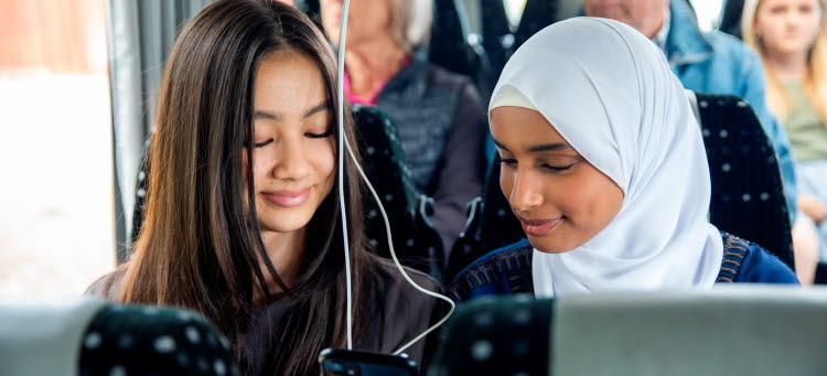 Två tjejer i tonåren sitter i en buss och tittar på en mobiltelefon. 