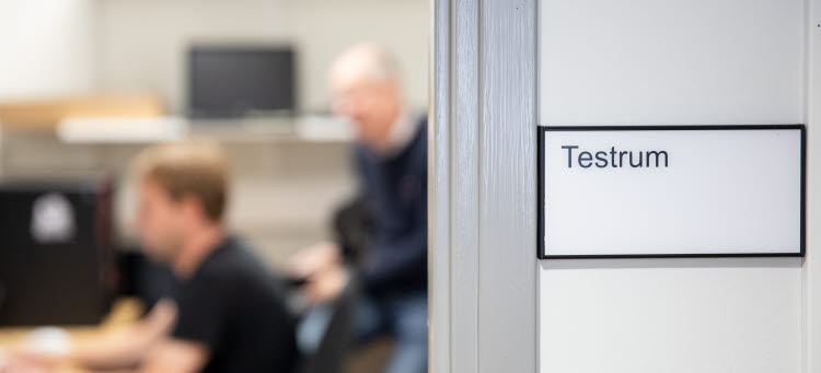 Två personer i ofokus som sitter i ett rum med en skylt utanför där det står "Testrum".