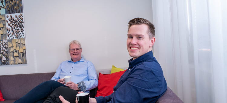 Två personer sitter i en soffa. De ler båda in i kameran.