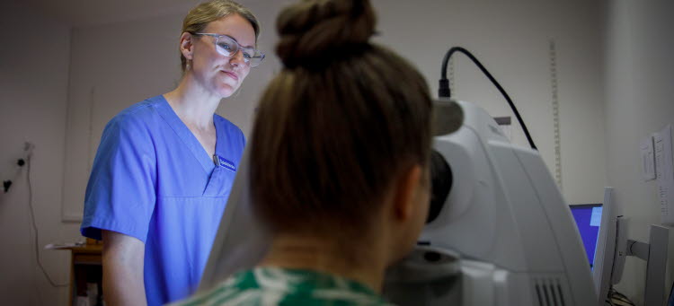 En patient undersöker sina ögon genom att titta in en maskin. Sjuksköterskan Therés Fransson övervakar. 